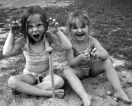 Two young girls girls sitting on the ground, one making making a snarling face with hands arced into claws.  The other, laughing.